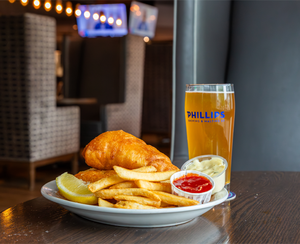 Fish and chips on a plate with a pint of phillips beer sitting on a dark wooden table