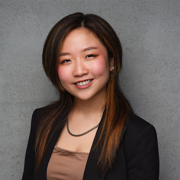 Person with dark hair a blazer standing in front of a grey background.
